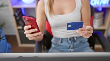 Genç bir kadın, mavi ışıkla aydınlatılmış modern bir oyun odasında akıllı telefon ve kredi kartı tutuyor..