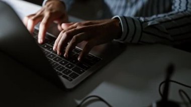 Karanlık bir ofis ortamında dizüstü bilgisayarda yazı yazan bir adamın ellerinin yakın çekimi, iş ve teknoloji tasviri.