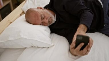 Yatak odasında sakallı, akıllı telefon tutan, uykusuzluğu ya da sabah rutinini yansıtan olgun kel bir adam..