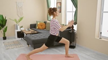 Genç bir kadın oturma odasında yoga yapıyor, sağlığını ve ev egzersizlerini modern bir apartman ortamında resmediyor..