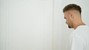 Profil görüntüsü: Sakallı, beyaz bir odada duran, düz beyaz tişörtlü genç bir adam..