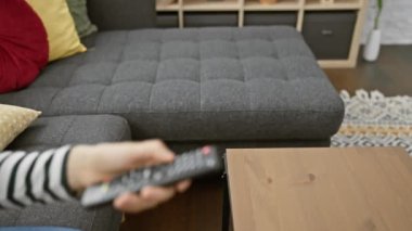Kanepesi ve ev dekoru olan modern bir oturma odasında uzaktan kumandayı elinde tutan kişi..