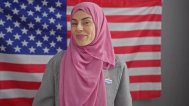 Pembe tesettürlü gülümseyen bir kadın Amerikan bayrağının önündeki kamerayı işaret ediyor, çeşitliliği ve katılımı simgeliyor..