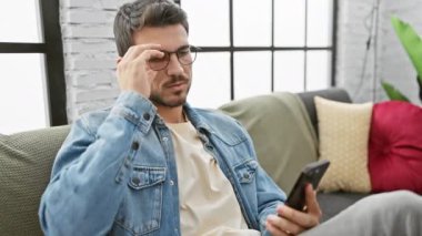 Modern bir oturma odasında akıllı telefonuna bakarken kendini stresli hisseden sakallı ve gözlüklü genç bir İspanyol..