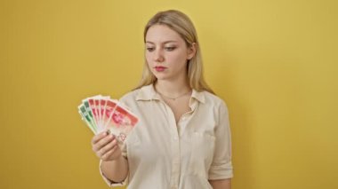 Kendine güvenen genç sarışın kadın seni İsrail banknotlarıyla işaret ediyor. İzole edilmiş sarı arka planda ciddi bir ön kamera görüntüsü.