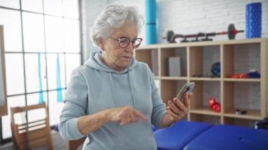 Fizyoterapi kliniğinin rehabilitasyon odasında akıllı telefon kullanan kıdemli bir kadın..