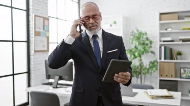 Modern bir ofiste tablet ve telefon kullanan, takım elbiseli, sakallı ve gözlüklü sofistike bir orta yaşlı adam..