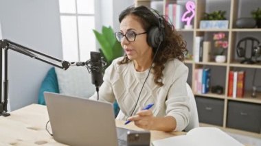 İspanyol kadın radyo stüdyosunda mikrofon ve dizüstü bilgisayarla ahşap masada podcast yapıyor.