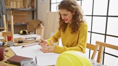 Stresli genç kadın, eli başının üstünde, marangozluk masasında kağıt üzerine çizerken öfkeyle hayal kırıklığını ifade ediyor. Şaşırmış ve güzel bir şekilde oturuyor, stüdyonun endişeli atmosferini somutlaştırıyor..