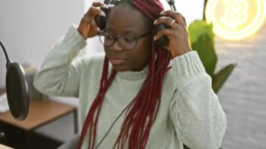 Örgülü Afrikalı bir kadın radyo stüdyosunda kayıt yapıyor, mikrofon önünde kulaklık takıyor..