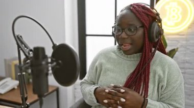 Örgü örgülü Afrikalı Amerikalı bir kadın, Bitcoin neon levhası olan bir stüdyoda podcast yapıyor..
