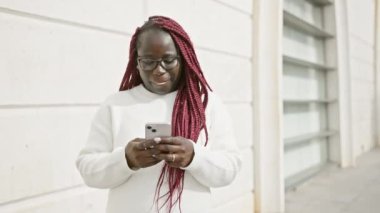 Saçları örgülü ve gözlüklü gülümseyen Afrikalı kadın şehir caddesinde akıllı telefon kullanıyor..