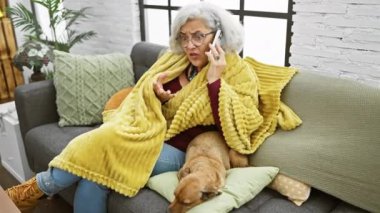 Olgun bir kadın telefonda konuşuyor, endişeli bir ifade, evcil köpekle evde, rahat bir ev ortamında..