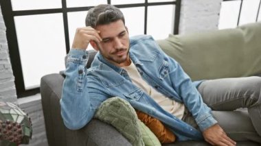 Kot ceketli yakışıklı bir İspanyol, modern bir oturma odasındaki gri bir koltukta düşünceli bir şekilde oturur..