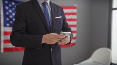 Takım elbiseli profesyonel İspanyol adam arka planda Amerikan bayrağı olan bir akıllı telefon kullanıyor..
