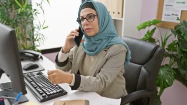 Ofis masasında çalışan, içeride telefonla konuşan, tesettürlü orta yaşlı bir kadın.