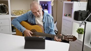 Kel, olgun bir adam modern bir ofiste gitar çalıyor. Arka planda tablet ve müzik aletleri var..