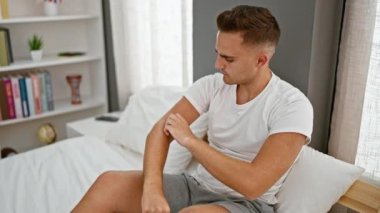 Düşünceli bir genç adam, iyi aydınlatılmış bir yatak odasında oturuyor, rahat giyinmiş, düşünceli bir ruh hali içinde..