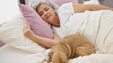 Gri saçlı olgun bir kadın, rahat bir yatak odasında kahverengi köpeğiyle birlikte huzur içinde uyuyor..