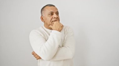 Düşünceli orta yaşlı Latin beyefendi, süveter giyiyor, beyaz arka planda soyutlanmış duruyor, çenesi elinin üzerinde dinleniyor ciddi bir soru üzerine kafa yorarken..