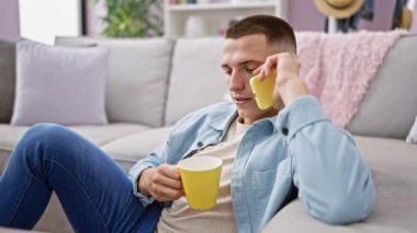 Rahat genç adam, rahat oturma odasında akıllı telefonla konuşurken elinde kahve fincanı tutuyor.