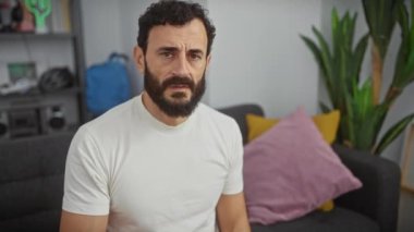 Stresli orta yaşlı bir adam, elleri alnında dinleniyor, evinde beyaz tişörtü ile endişeli, ezici bir krizle boğuşuyor, gözle görülür şekilde stresli ve sorunlu..