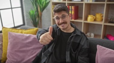 Neşeli genç İspanyol adam, gözlük takıyor, kendine güvenen bir jest yapıyor, evdeki kanepede oturuyor. Pozitif adamın mükemmel tasvip ifadesi, dostça bir portre için sevinçle gülümsemesi..