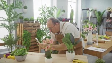 Gözlüklü ve kahverengi önlüklü yaşlı bir adam arka planda bir dizi bitkiyle birlikte parlak bir çiçekçide çiçekleri inceliyor..