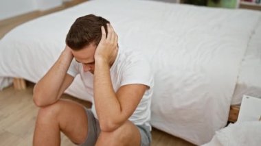 Sıkıntılı genç bir adam yatak odasındaki bir yatağın yanında oturmuş, endişe veya baş ağrısı tasvir ediyor..
