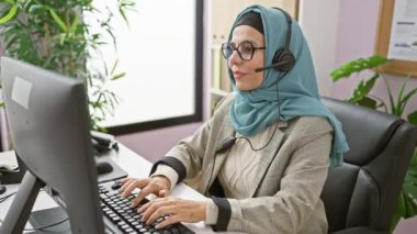 Modern bir ofiste tesettürlü ve gözlüklü olgun bir kadın masasında gülümsüyor.