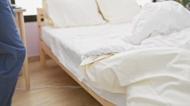 Gündelik giysiler içinde genç bir adam. Parlak, modern bir yatak odasında dağınık bir yatak topluyor. Sabah rutinini gösteriyor.