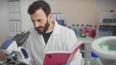 Laboratuvar önlüklü orta yaşlı sakallı bir adam elinde bir defter tutarken laboratuarda mikroskopla örnekleri analiz ediyor..