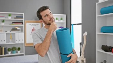 Genç İspanyol bir adam, sakallı, ciddi yüzünü çerçeveleyen, rehabilitasyon kliniğinde yoga minderini tutan, elleri çenesinde, ilginç bir şekilde kafa karıştırıcı bir fikri düşünen.