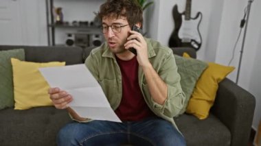 Gözlüklü İspanyol adam elinde belge tutarken telefonla konuşuyor, rahat bir oturma odasında oturuyor..