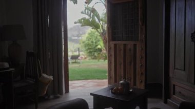 İçi açık ahşap kapılı, güneşli bir bahçeye açılan, sehpada kahve, ortam ve rahat mobilyalar..