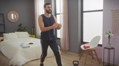 Günlük spor kıyafetleri içinde sakallı bir adam modern bir yatak odasında egzersiz yapıyor. Bu da evde spor yaptığını gösteriyor..