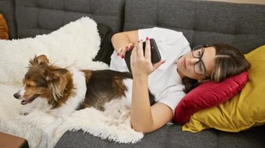 Genç bir kadın evinde akıllı bir telefonla köpeğiyle kanepede vakit geçirmekten zevk alır..