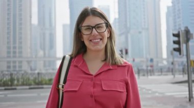 Pembe tişörtlü, gözlüklü, gülümseyen bir kadın modern Dubai marina siluetinin önünde duruyor ve şehir keşiflerini ve seyahatlerini anlatıyor..