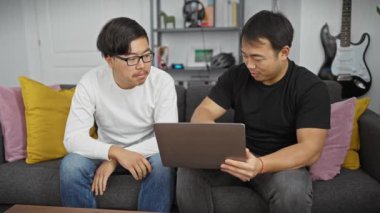 İki Asyalı, modern bir oturma odasında arka planda gitarlı bir dizüstü bilgisayar kullanıyor.