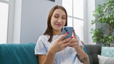 Neşeli genç bir kadın akıllı telefonunu evde rahat bir koltukta, rahat bir yaşam tarzını somutlaştırarak kullanmaktan hoşlanır..