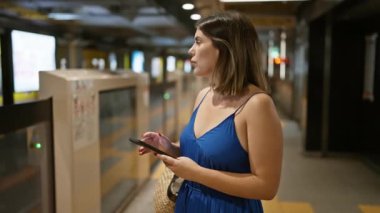 Neşeli, güzel İspanyol bir kadın, neşeli bir yolcu, metro istasyonunda metro trenini bekliyor, telefonunun ekranına dalmış, gülümseyen yolculuğuna dalmış.
