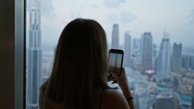 Bir kadın, alacakaranlıkta gözlemevinden akıllı telefonuyla Dubai siluetini ve gökdelenlerini yakalıyor..