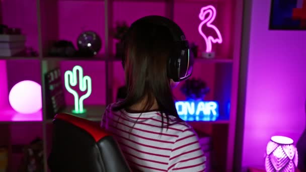 夜にネオンライトのゲームルームでヘッドフォンを着用する若いブルネットの女性は 現代の家庭のエンターテイメント環境を描いています — ストック動画