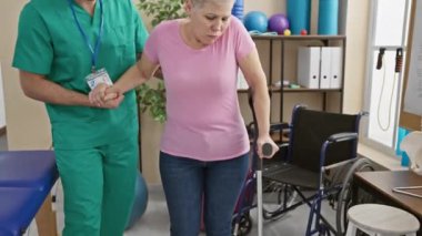 Bir erkek fizyoterapist, yürüyen bir kadına tekerlekli sandalyeyle klinikte yardım ediyor..