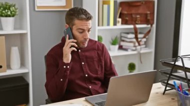 Bordo gömlekli, sakallı, odaklanmış genç bir İspanyol. Modern bir ofiste dizüstü bilgisayarla konuşurken akıllı bir telefonla konuşuyor.