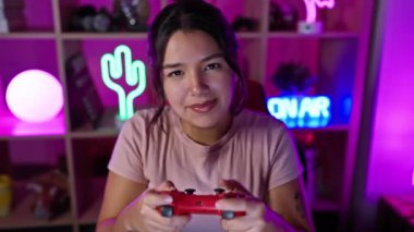 Genç bir İspanyol kadın, kırmızı bir oyun kontrolörüyle oynarken gülümsüyor. Canlı, neon ışıklı bir oyun odasında.
