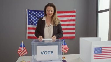 Kendine güvenen bir kadın, arka planda demokrasiyi temsil eden Amerikan bayraklarıyla bir Amerikan seçim merkezinde oy kullanıyor..