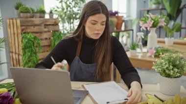 Laptopta çalışan ve çiçekçi dükkanında renkli çiçekler ve yeşil bitkilerle notlar alan İspanyol bir kadın..