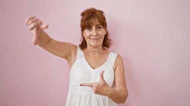 Elbiseli çekici orta yaşlı bir kadın, pembe arka planda elleri ile bir fotoğraf fikrini çerçeveletiyor. Yaratıcılığını serbest bırakarak mutluluk ve ilhamla ışıldıyor..