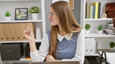 Neşeli genç sarışın iş kadını kendine güvenerek çeşitli yönleri işaret ediyor, ofiste çekici gülüşünü gösteriyor. Reklamınız için mükemmel kopyalama alanı!